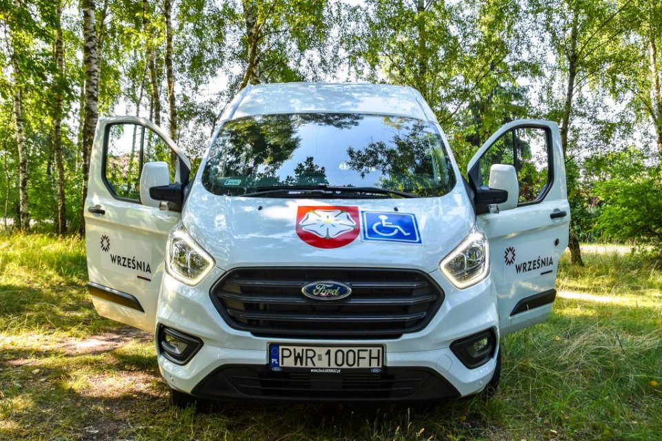 Biały bus marki ford przedstawiony od frontu na tle drzew. Na masce pojazdu widoczny herb Gminy Września oraz symbol niepełnosprawności.