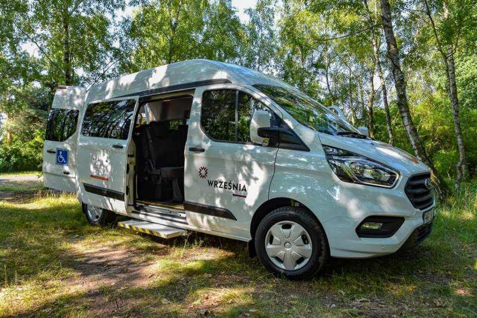 Biały bus przedstawiony od boku na tle drzew. Drzwi busa uchylone, widoczna rampa dla osób niepełnosprawnych. Na karoserii umieszczony symbol niepełnosprawności oraz logo Gminy Września.